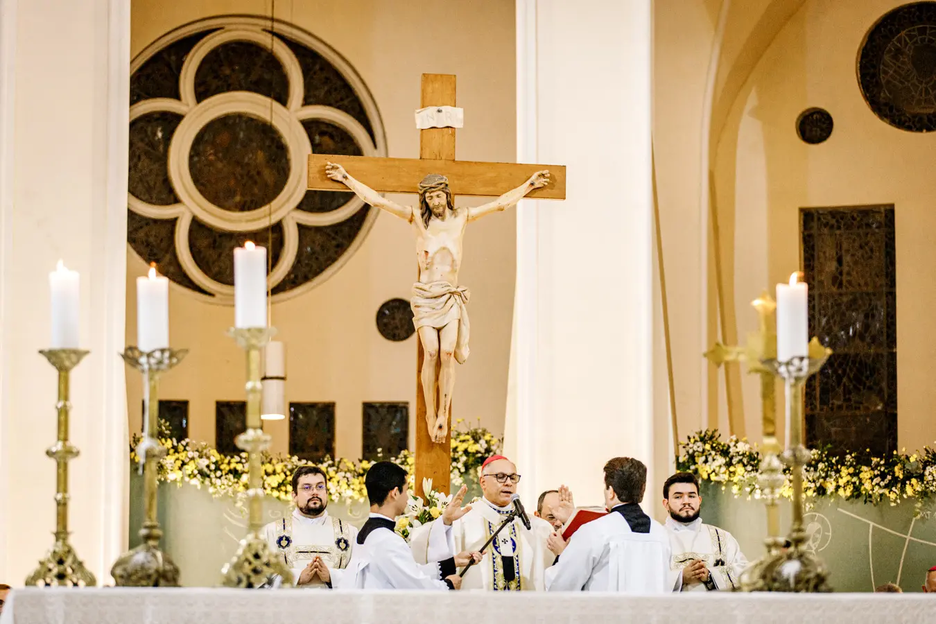 Cerimônia religiosa em uma igreja, com um bispo celebrando a missa diante de um grande crucifixo com a imagem de Jesus crucificado, cercado por sacerdotes, coroinhas, velas acesas e arranjos de flores no altar.