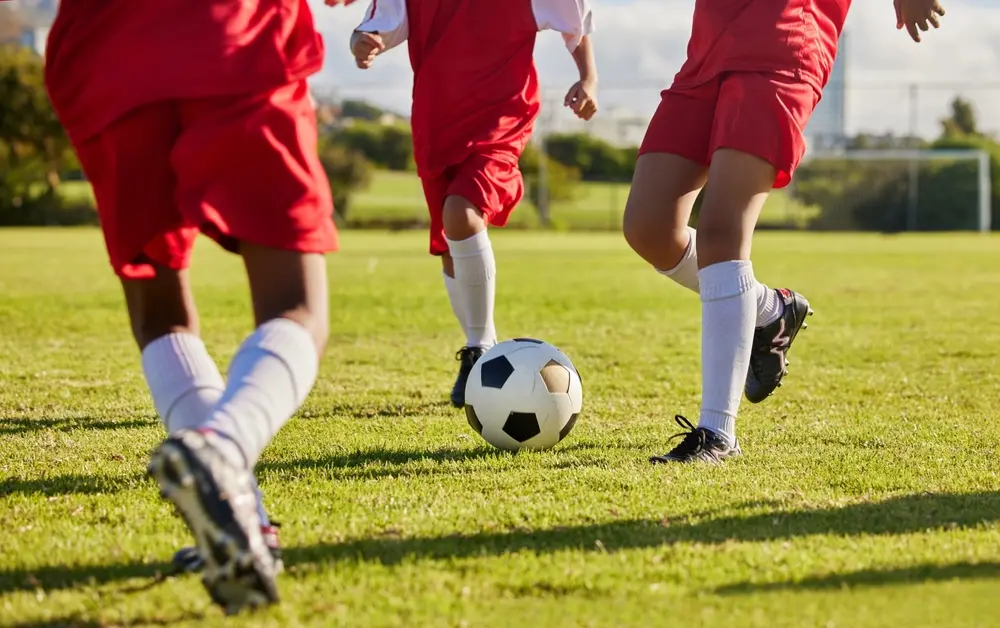 Crianças com uniformes vermelhos disputam a bola durante uma partida de futebol em um campo gramado, usando meiões brancos e chuteiras, em um dia ensolarado.