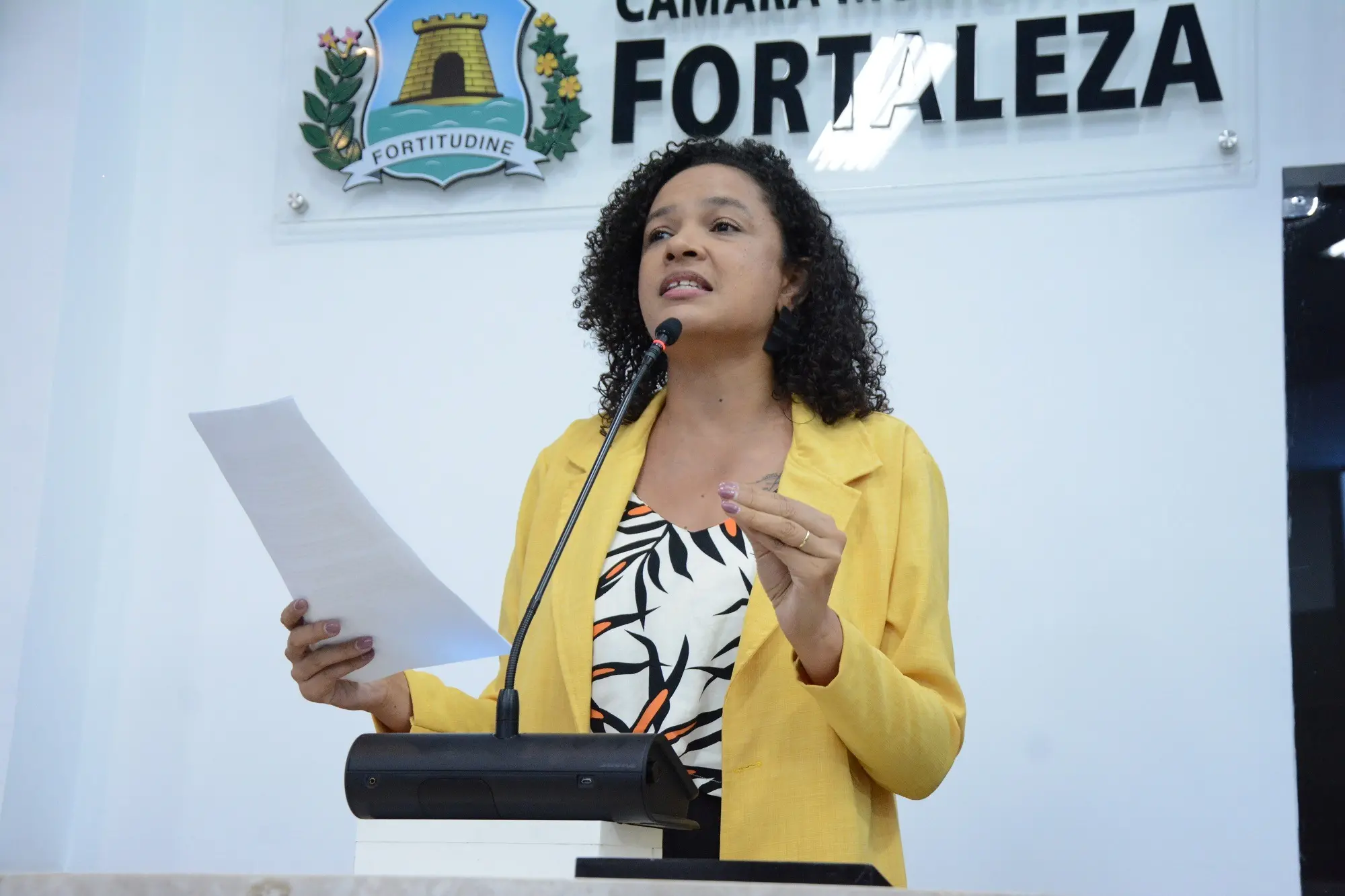 Mulher falando em um púlpito durante sessão na Câmara Municipal de Fortaleza, segurando um papel e discursando com microfone, com banner “Câmara Municipal de Fortaleza” ao fundo.