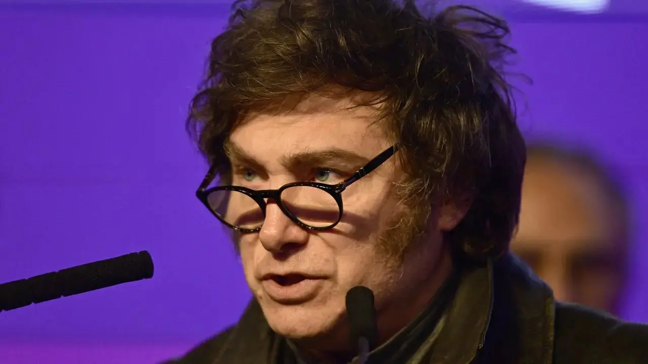 Homem com cabelo castanho bagunçado e óculos de aro preto, falando em um palco com microfone, em ambiente iluminado com fundo azul e roxo