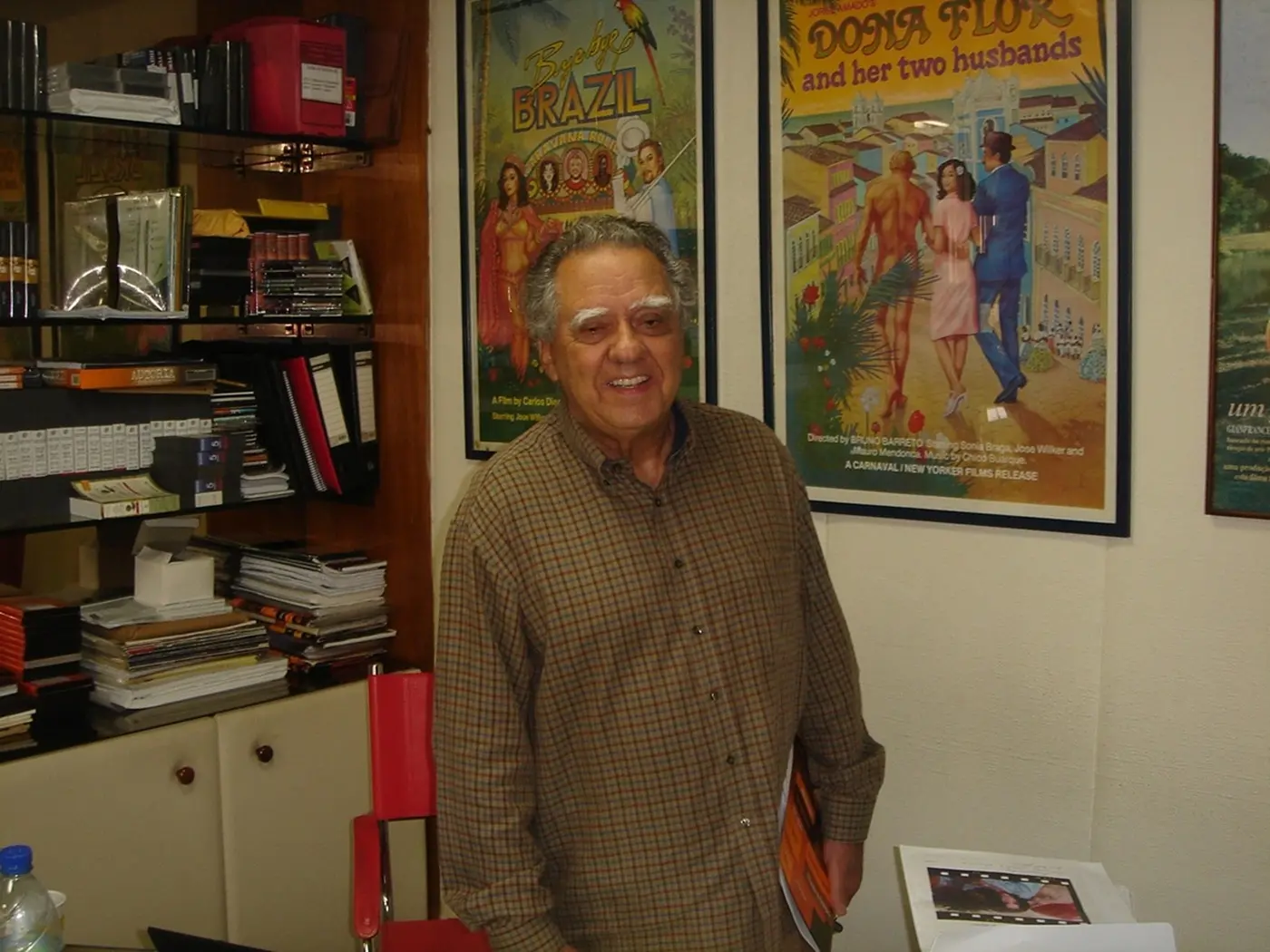 O cineasta Luiz Carlos Barreto sorri em pé no centro da imagem, vestindo uma camisa xadrez de mangas compridas em tons de marrom e bege. Ao fundo, destacam-se pôsteres emoldurados dos filmes 'Bye Bye Brasil' e 'Dona Flor e Seus Dois Maridos', além de prateleiras com livros e documentos.