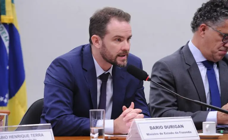 Dario Durigan sentado à mesa durante uma audiência ou reunião oficial, falando ao microfone. A pessoa veste terno azul-marinho, camisa clara e gravata escura, com as mãos apoiadas sobre a mesa. À frente há um copo com água e uma placa de identificação com o texto “DARIO DURIGAN” e “Ministro de Estado da Fazenda”.
