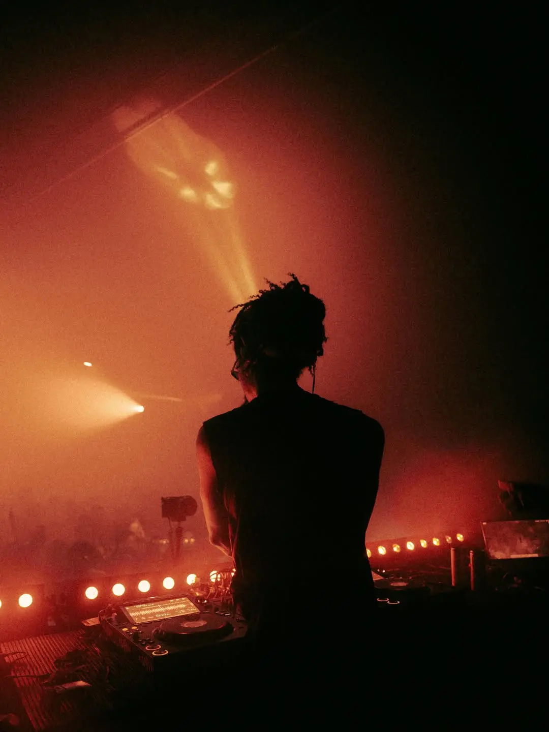 Foto da silhueta de um DJ de costas, mexendo na mesa de mixagem. Ao redor dele, luzes vermelhas iluminam a imagem.