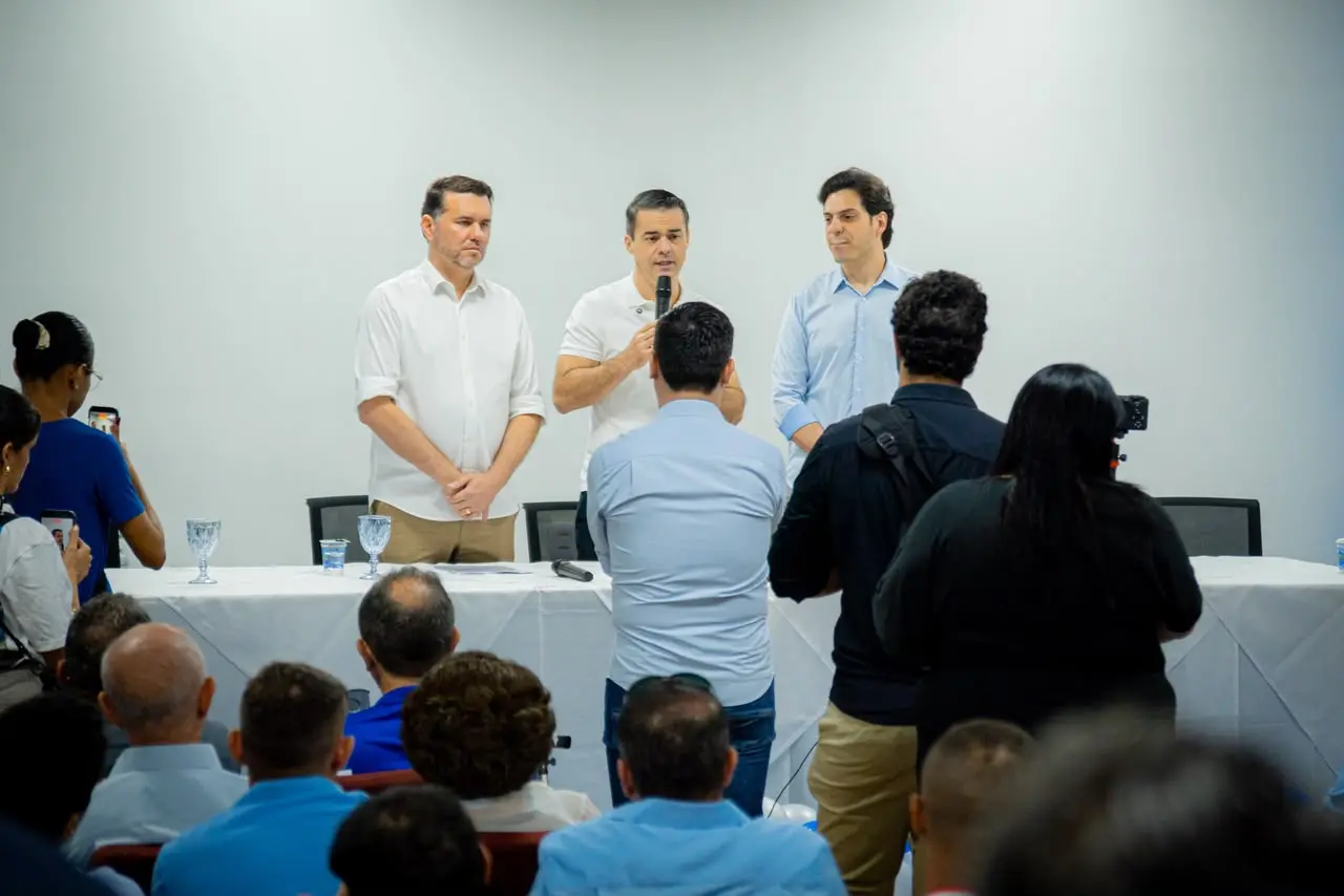 Três homens estão à frente de uma mesa durante um evento. O homem ao centro fala ao microfone, enquanto os outros dois permanecem ao lado, ouvindo. À frente deles, jornalistas e participantes registram o momento com celulares e câmeras. O auditório está ocupado por pessoas sentadas, voltadas para a mesa, em um ambiente de paredes claras e iluminação branca.