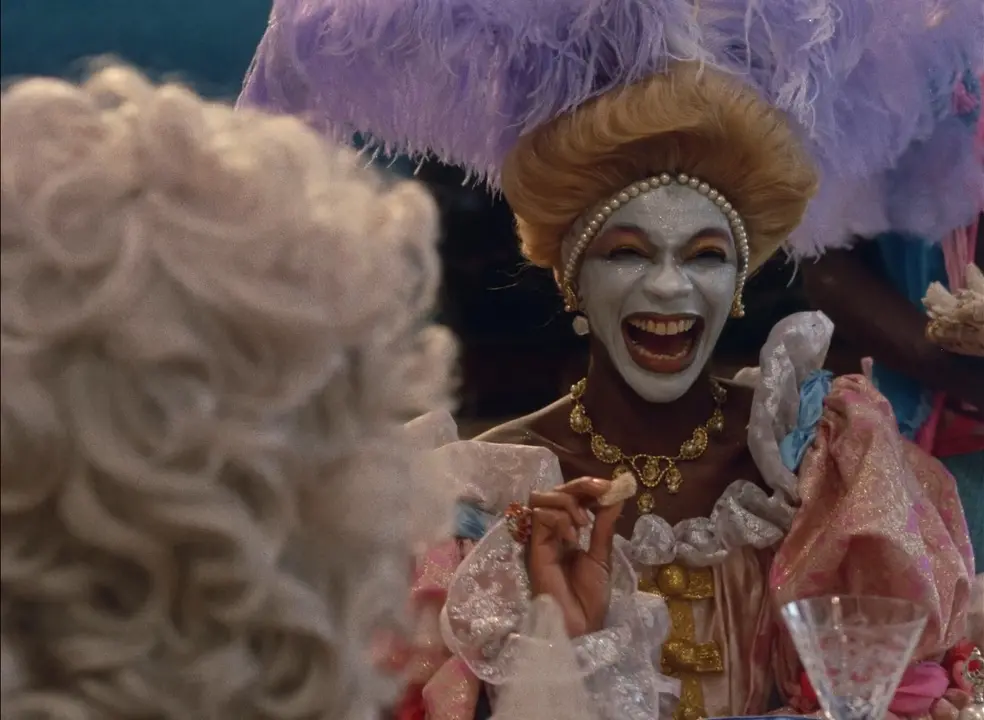 Imagem de divulgação do filme 'Xica da Silva', com Zezé Motta, mostra uma mulher negra com o rosto maquiado de branco ri ruidosamente enquanto segura um alimento entre os dedos, vestindo figurino de época suntuoso e penteado ornamentado com pérolas e penas lilases. No primeiro plano, de costas e desfocada, vê-se a cabeça de outra pessoa com peruca cacheada clara.