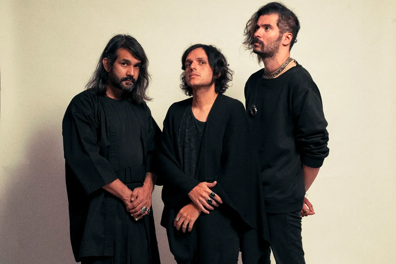 Um trio de homens brancos posa para a foto. Os três estão com roupas compridas, totalmente pretas, e alguns acessórios de prata.