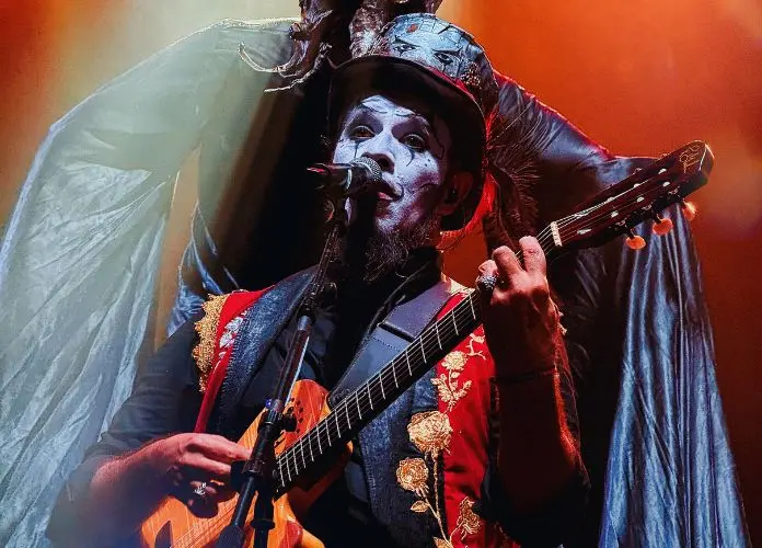 Foto de um homem com pintura facial, cartola e vestes coloridas tocando guitarra.