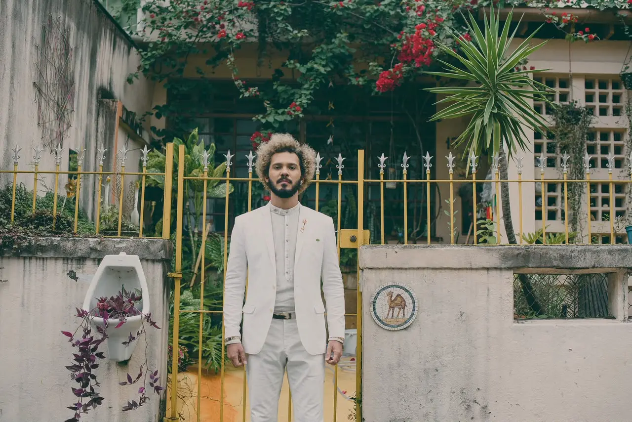 Um homem de terno e barba está em pé diante de um portão. Atrás do portão há plantas, flores, uma fachada de casa e um muro com mosaico.