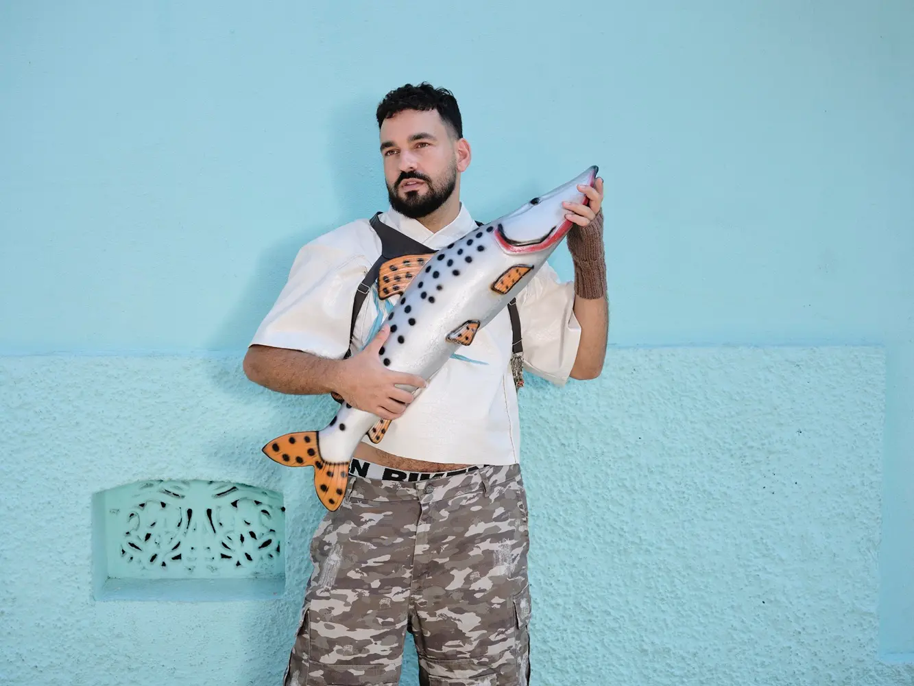Um homem de barba segura uma escultura de peixe com as mãos diante de uma parede. Ele veste camisa, calça de camuflagem e olha para o lado.