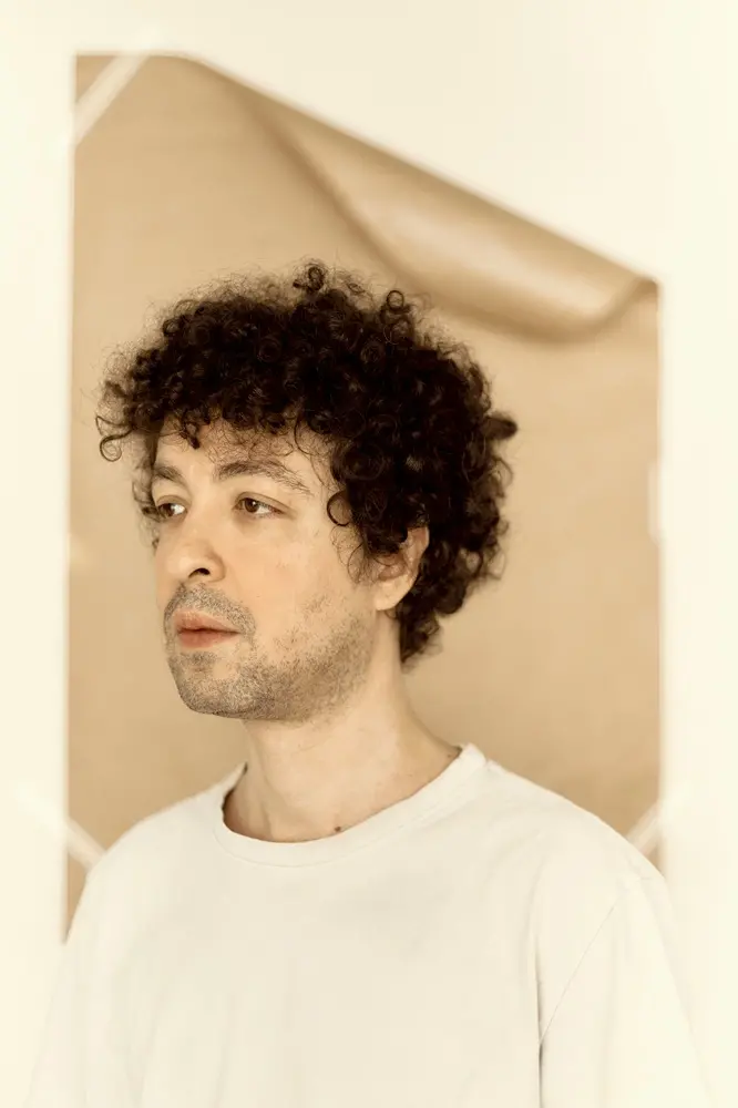 Um homem branco, de cabelo cacheado e barba, veste uma camiseta de cor clara. Ele olha para o lado diante de um fundo de papel.