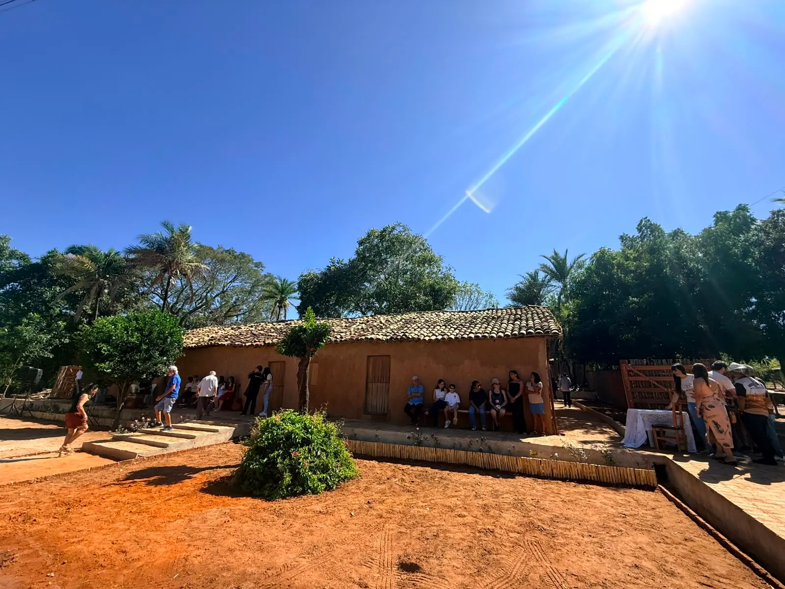 Uma fotografia ao ar livre sob um céu azul brilhante e sol intenso mostra uma casa de taipa com telhado de cerâmica. Várias pessoas estão reunidas e sentadas ao longo da base da casa, que tem paredes de barro e portas de madeira. O chão à frente é de terra batida vermelha. Árvores e palmeiras cercam a propriedade.