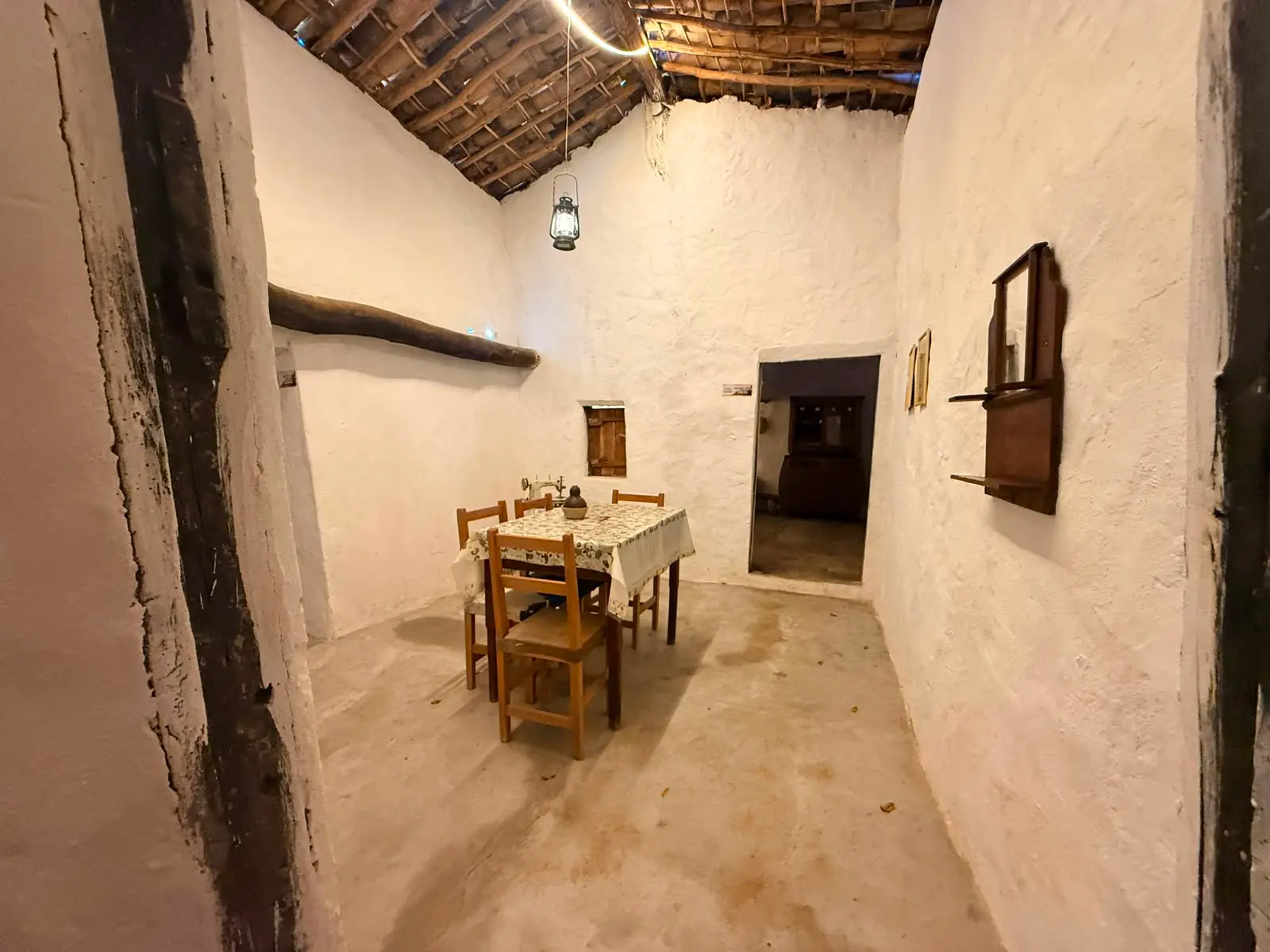 Uma vista interna de uma sala de jantar simples dentro de uma casa de taipa com paredes de barro caiadas de branco e um telhado de madeira e palha exposto. Uma mesa de madeira com uma toalha de renda e quatro cadeiras está no centro. Uma lanterna a querosene está pendurada no telhado. Há uma pequena janela de madeira e um oratório na parede.