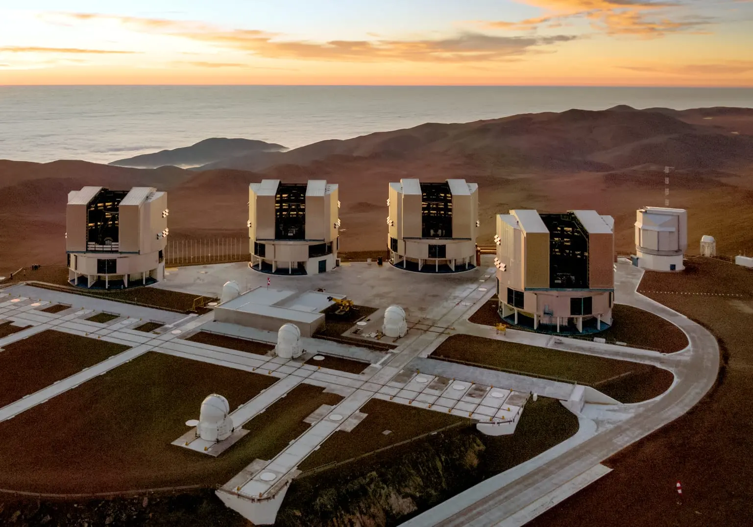 Vista aérea de um complexo astronômico no topo de uma montanha árida durante o pôr do sol. Destacam-se quatro grandes estruturas telescópicas modernas alinhadas em uma plataforma, cercadas por domos menores e um mar de nuvens ao fundo sob luz alaranjada.