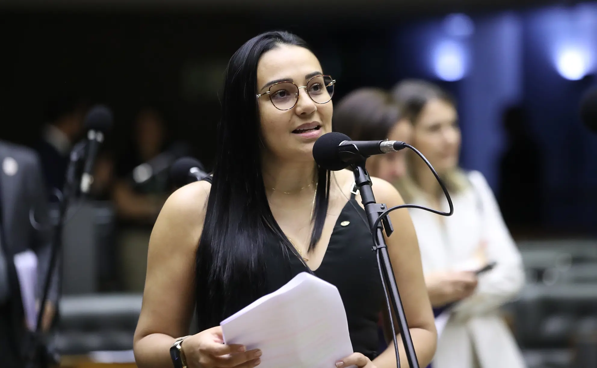 Uma mulher de pele negra e cabelos pretos longos, usando óculos de armação fina e blusa preta, fala ao microfone enquanto segura folhas de papel em um ambiente formal de plenário da Câmara dos Deputados.