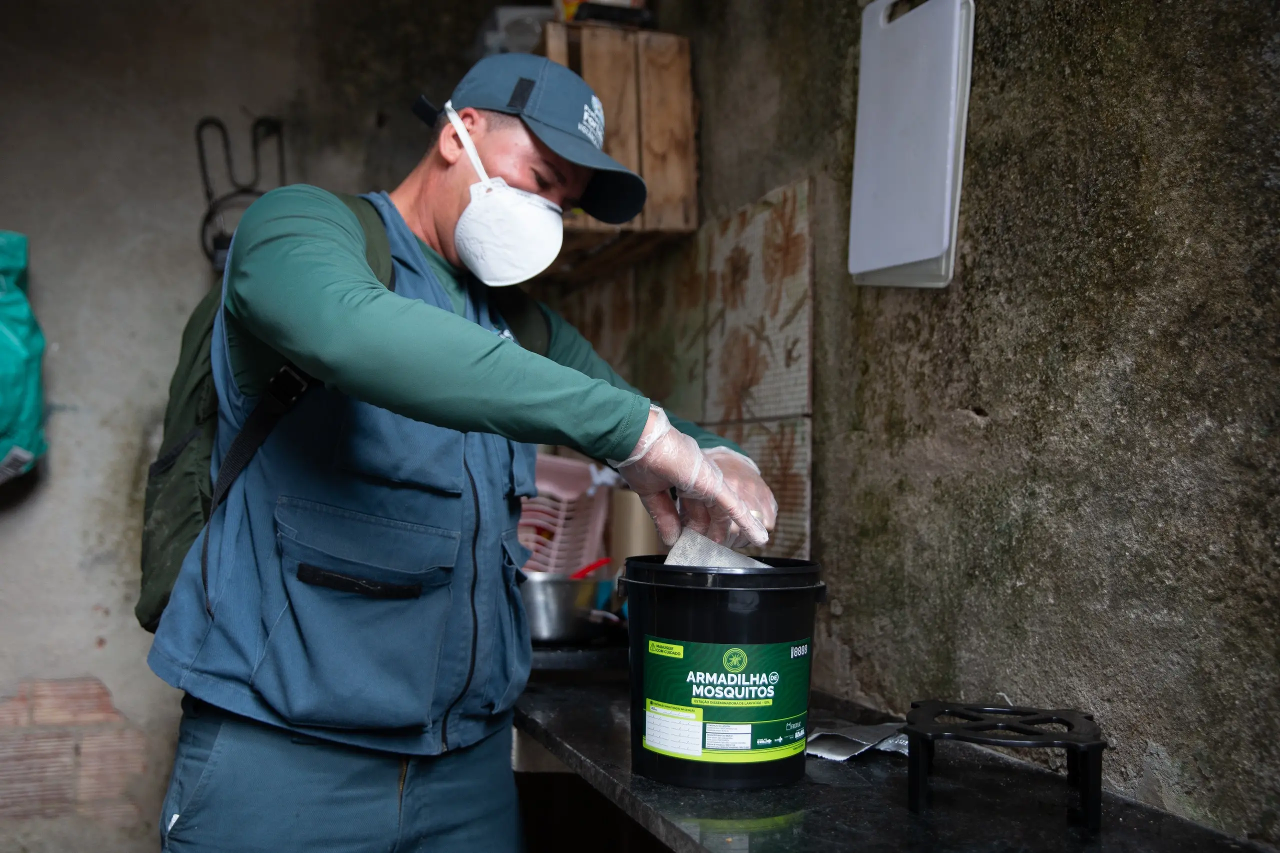 Agente de Endemias uniformizado, com máscara e luvas, instalando Armadilhas de Mosquitos, as Estação Disseminadora de Larvicida, em ambientes internos de uma residência para combater o Aedes aegypti
