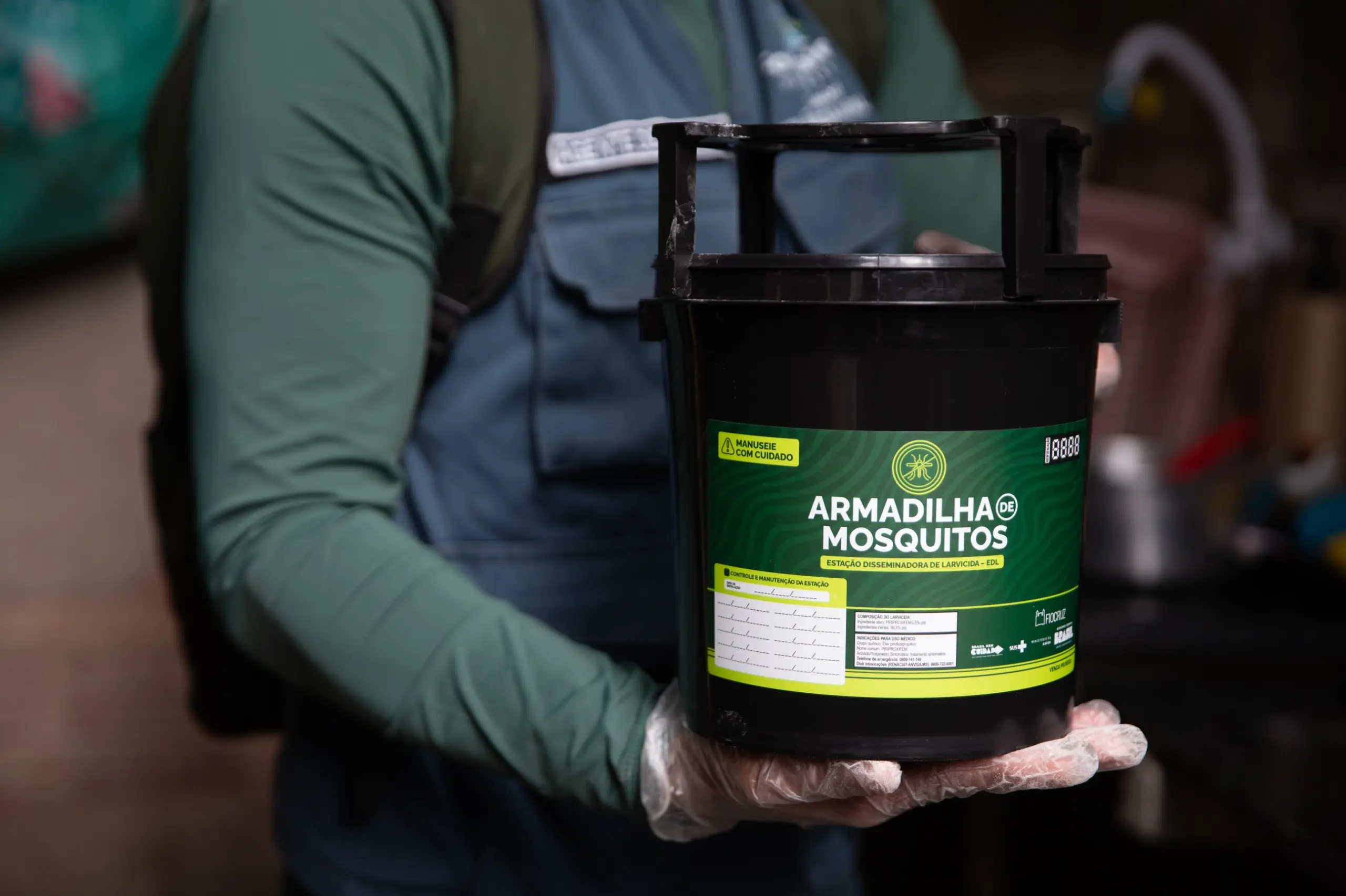 Agente de Endemias uniformizado, com máscara e luvas, instalando Armadilhas de Mosquitos, as Estação Disseminadora de Larvicida, em ambientes internos de uma residência para combater o Aedes aegypti