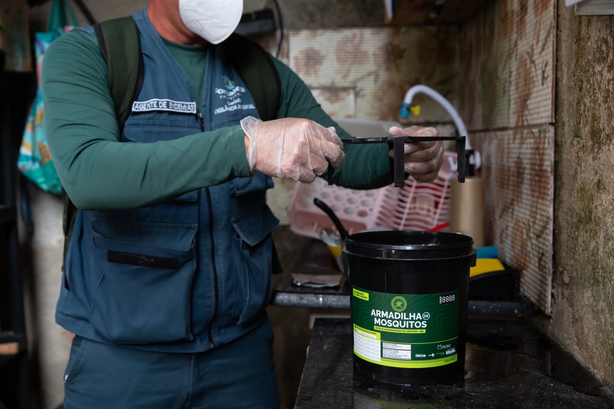 Agente de Endemias uniformizado, com máscara e luvas, instalando Armadilhas de Mosquitos, as Estação Disseminadora de Larvicida, em ambientes internos de uma residência para combater o Aedes aegypti