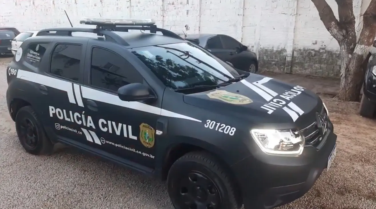 Viatura da Polícia Civil estacionada na rua, com faixas brancas e cinzas, logotipos oficiais e número 301208 no capô, em frente a um muro com manchas e árvore.
