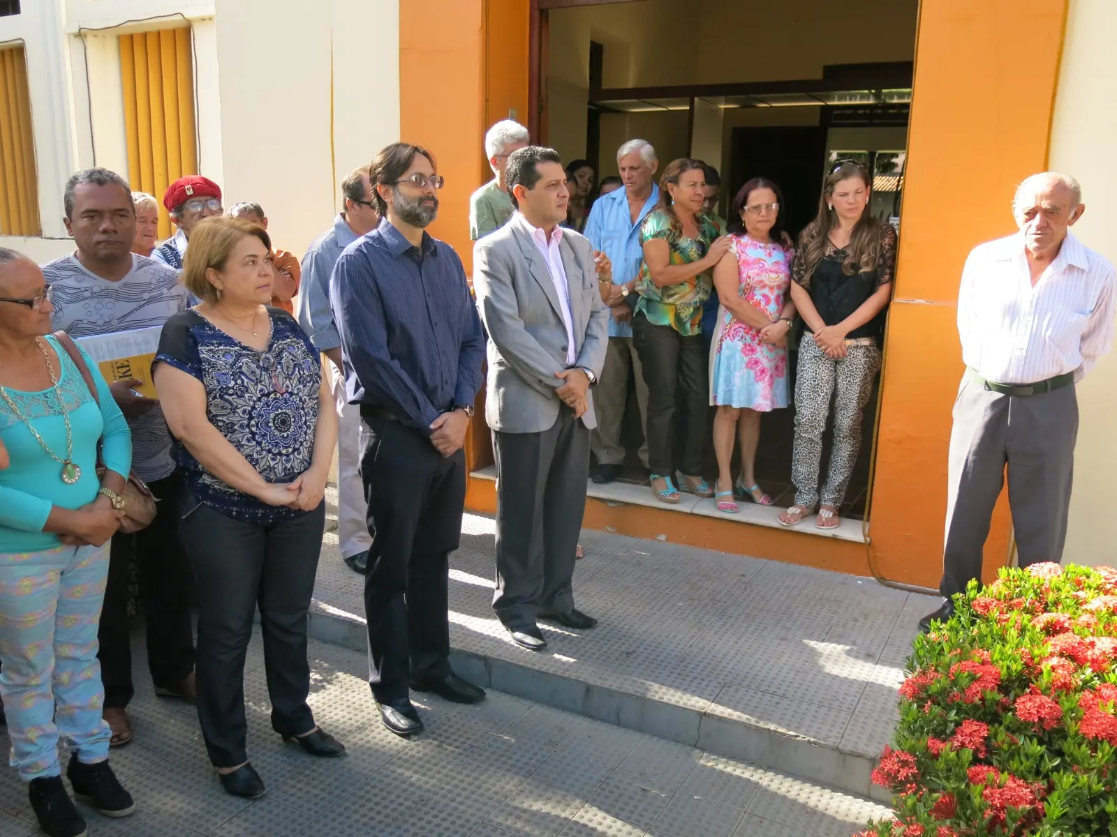 Grupo de pessoas reunidas na entrada de um prédio público com paredes amarelas e laranjas. À direita, um homem idoso de camisa social listrada de mangas compridas e calça cinza observa o evento de pé ao lado de um arbusto com flores vermelhas.