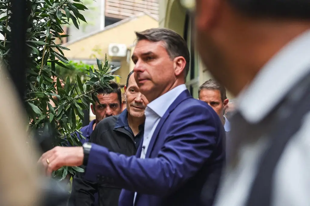 Pessoa de terno azul caminha por uma área externa cercada por outras pessoas, entre plantas e construções ao fundo. A imagem foi registrada de perto, com alguns elementos desfocados em primeiro plano, destacando o grupo em um corredor estreito ao ar livre.