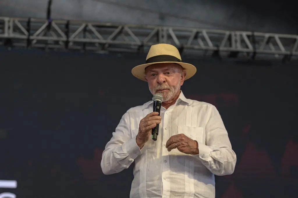 Homem idoso com chapéu claro e camisa branca segura um microfone no palco, falando durante um evento ao ar livre.