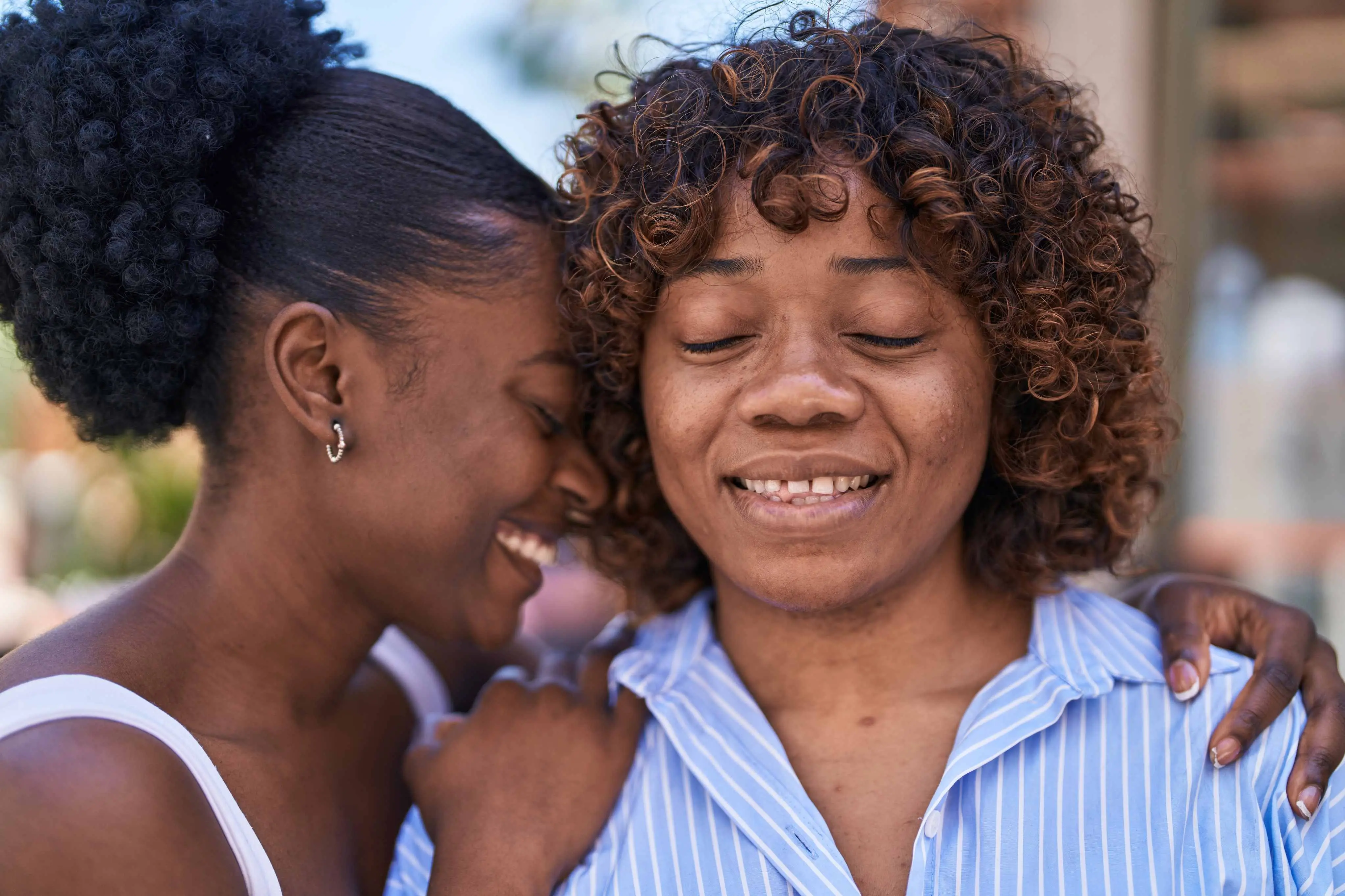 Fotografia em plano médio de duas mulheres negras expressando afeto. A mulher à direita está em primeiro plano, vestindo uma camisa de botões azul-clara com listras verticais brancas. Ela tem cabelos cacheados curtos em tom castanho com luzes mel, olhos fechados e um sorriso tranquilo. A mulher à esquerda está de perfil, vestindo uma regata branca e com o cabelo crespo preso em um penteado alto. Ela abraça a outra mulher por trás, apoiando as mãos nos ombros dela e encostando a testa na lateral de sua cabeça com um sorriso radiante. O fundo está suavemente desfocado.