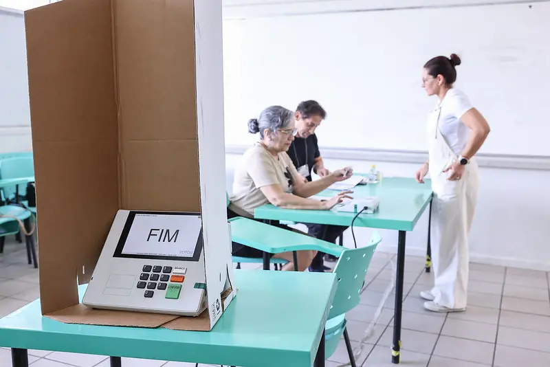 Cabine de votação em sala de aula escolar, com urna eletrônica dentro, contendo dígitos de 0 a 9 e botões 