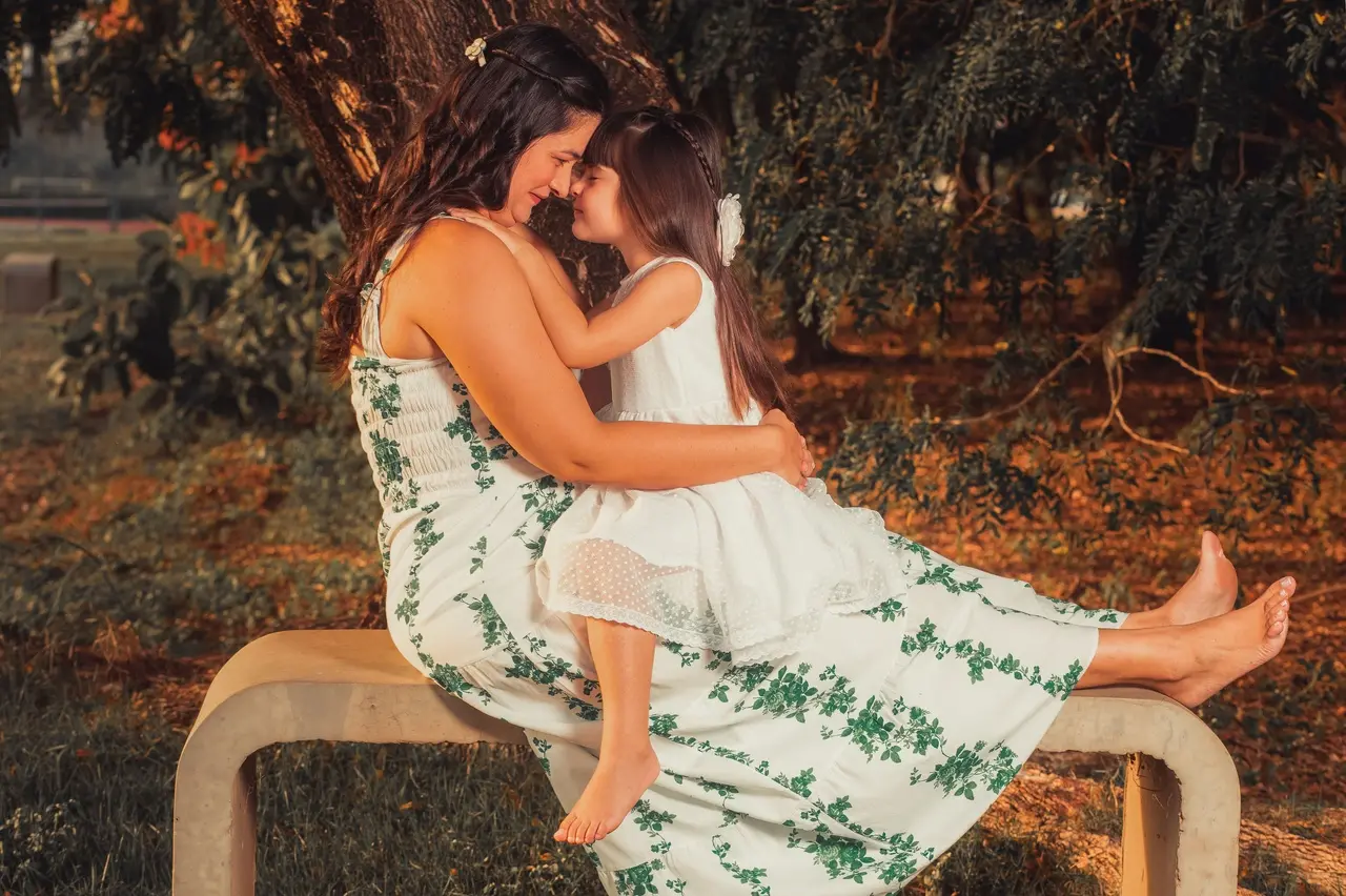 Mãe e filha se abraçam e encostam os rostos em um momento de carinho, sentadas em um banco em meio à natureza.