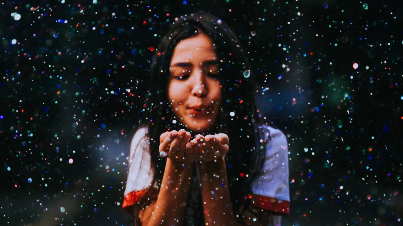 Uma jovem de cabelos castanhos sopra confetes coloridos que segurava nas suas mãos em concha.