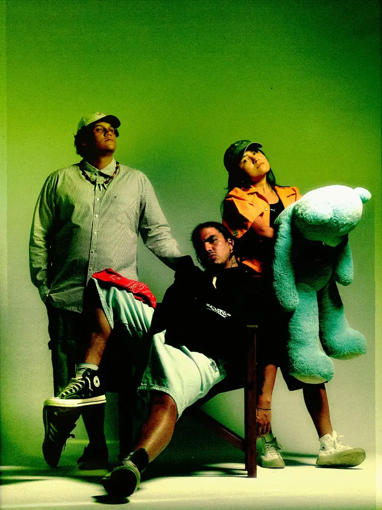 O trio Jambu posa para foto em frente a um fundo verde. São dois homens e uma mulher, que segura um urso de pelúcia azul.
