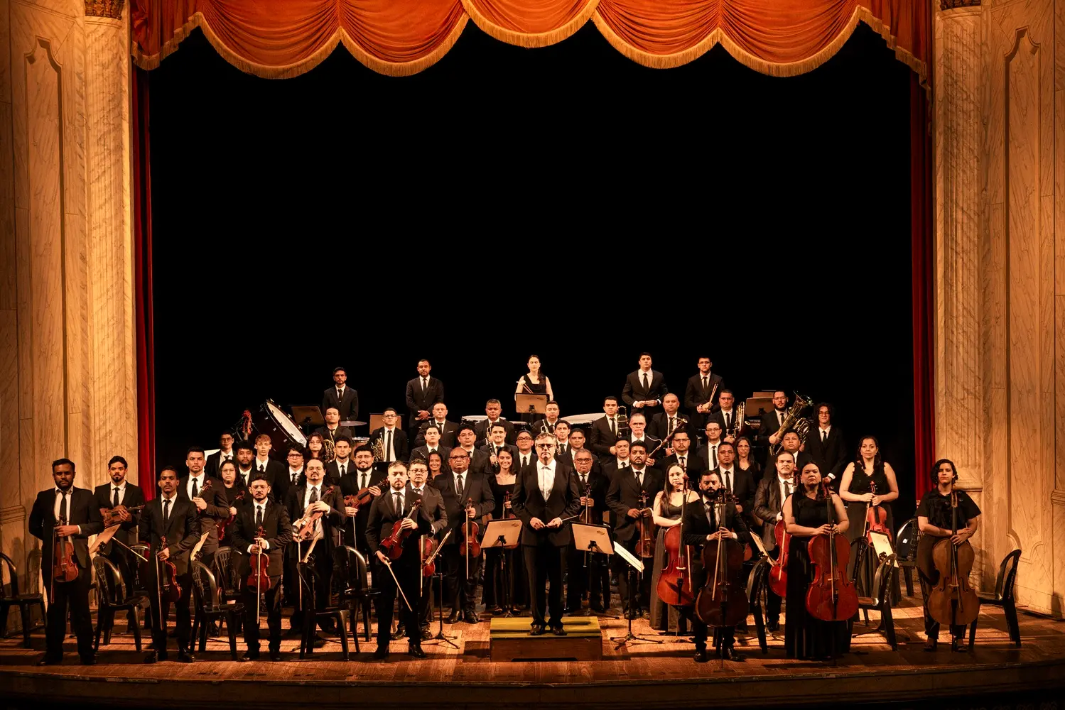 Foto mostra dezenas de músicos da Orquestra com seus instrumentos em um palco de teatro.