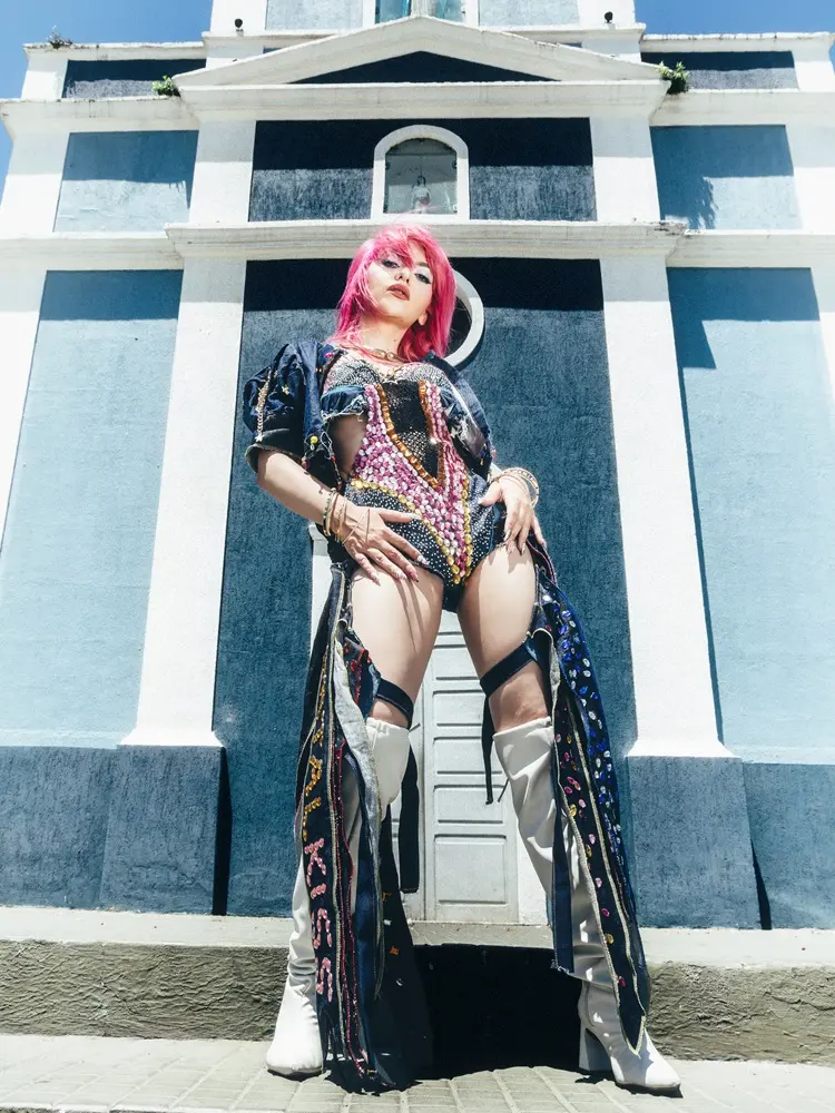 Cantora posa em frente a uma igreja azul. Ela tem cabelo rosa, maquiagem colorida, é branca e usa figurino colorido.