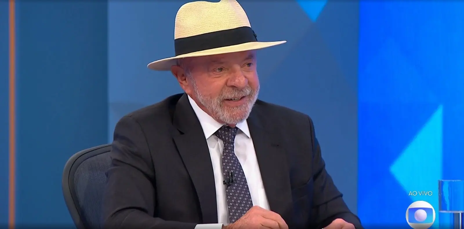 Presidente Lula, candidato na eleições 2026, aparece sentado, usa chapeu panamá, terno preto, gravata escura com estampa cinza e blusa branca.