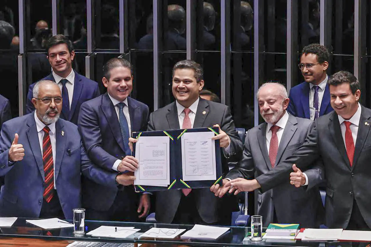 Grupo de autoridades, incluindo Presidente do Congresso Nacional David Alcolumbre e Presidente da República Lula (PT), em sessão oficial segura um documento aberto para exibição diante de uma mesa de trabalho. Ao redor, outras pessoas vestem ternos acompanham o momento. O ambiente possui painéis escuros ao fundo, copos com água e papéis sobre a mesa.