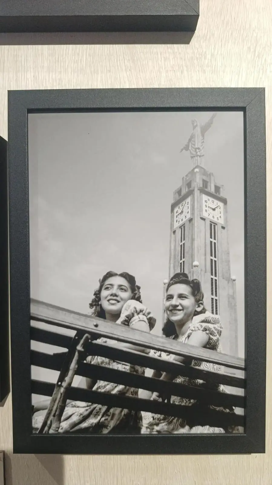 Fotografia em preto e branco de duas mulheres jovens sentadas em um banco com igreja ao fundo.