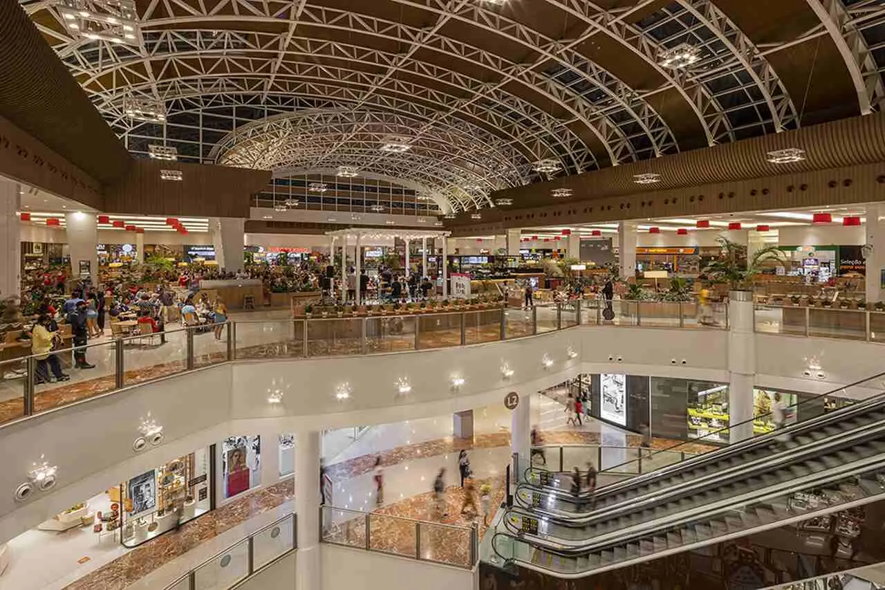 Fotografia do interior amplo e moderno de um centro comercial de vários pisos. Em primeiro plano, no lado direito, veem-se escadas rolantes que ligam os diferentes níveis do edifício.