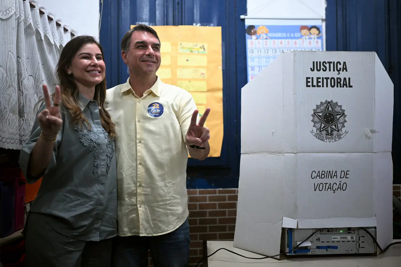 Cabina de votação da Justiça Eleitoral posicionada sobre uma mesa em local de votação. Ao lado da estrutura, duas pessoas posam para fotografia fazendo gesto com dois dedos erguidos. Ao fundo, há cartazes escolares afixados na parede, uma janela azul e uma tabela numérica pendurada. Na cabina, estão visíveis os textos 'JUSTIÇA ELEITORAL' e 'CABINA DE VOTAÇÃO', além do brasão da República.