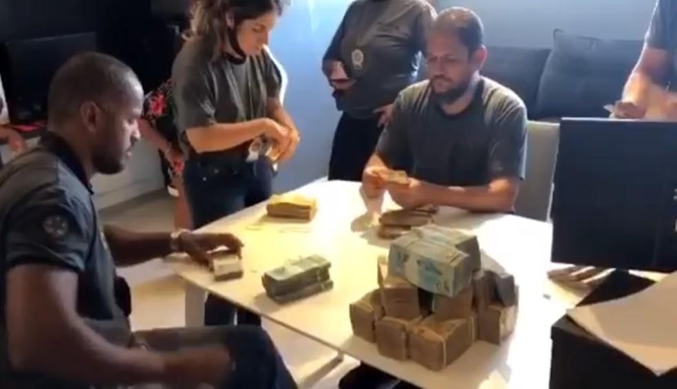Esta imagem mostra quatro agentes da Polícia Civil sentados em uma mesa branca contando cédulas de dinheiro encontradas na mansão do cantor Nego do Borel
