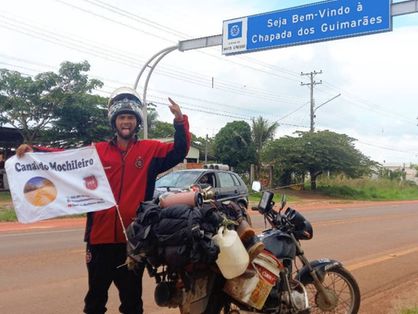 Tiago Bohrer em frente à moto e a uma placa da Chapada dos Guimarães, segurando bandeira do seu canal