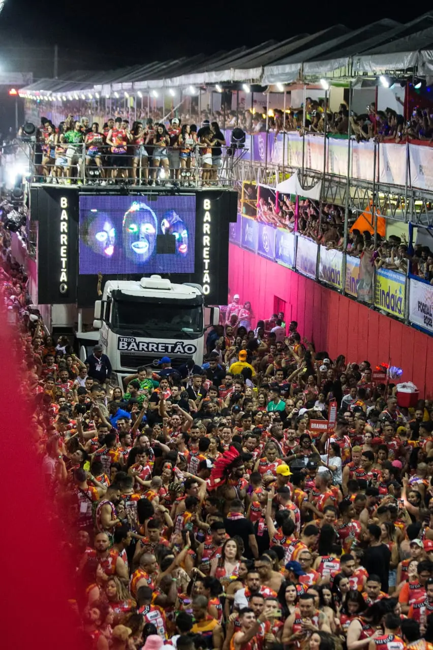 Imagem em plano aberto do corredor da folia lotado de foliões e o trio elétrico atravessando a região.