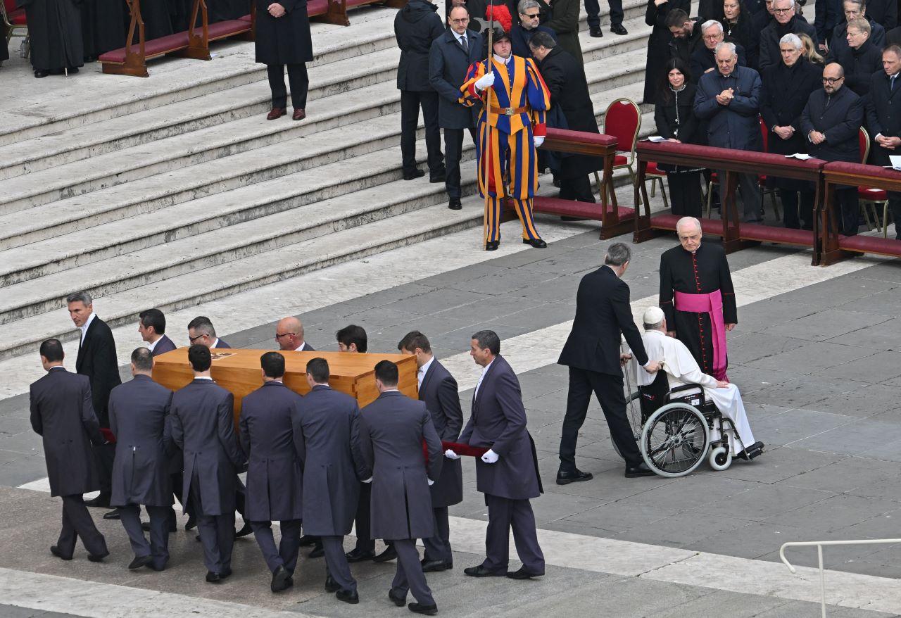 caixão é carregado por homens, do lado esquerdo da imagem, enquanto papa é transportado em cadeira de rodas