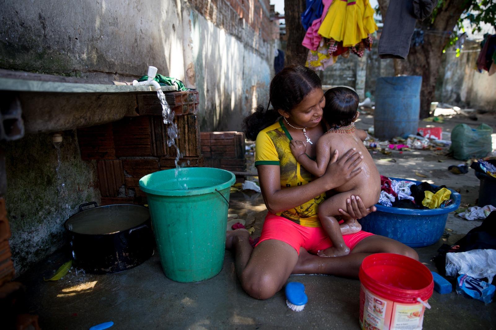 Mulher indígena venezuelana warao tomando banho com a filha perto de uma pia com água corrente e um varal com roupa colorida