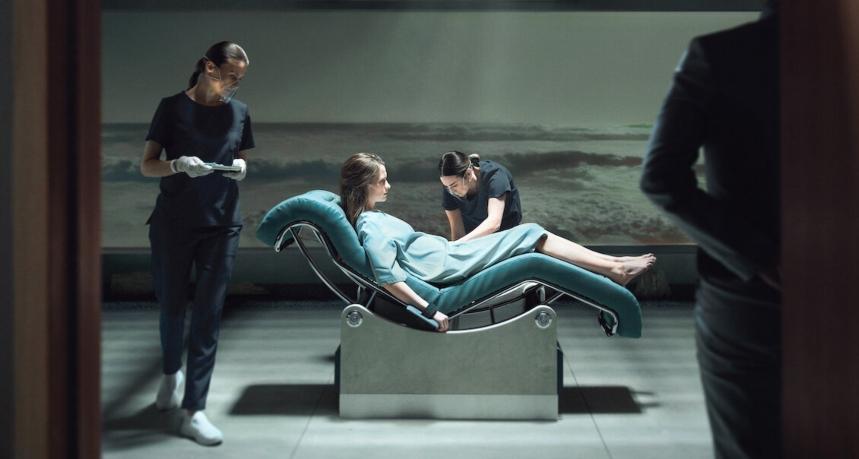 Numa sala, mulher jovem com roupa de hospital, fica deitada em uma cadeira e é atendida por duas jovens enfermeiras