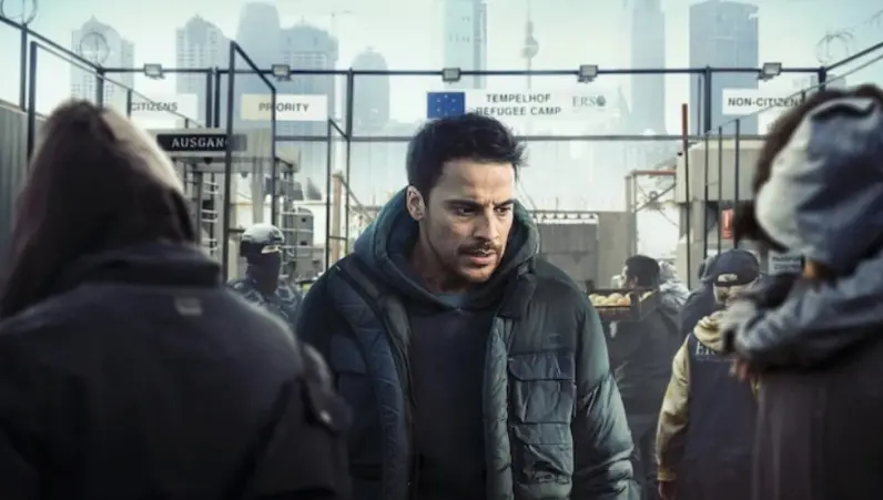 Homem jovem, de barba, vestindo roupa e casaco de inverno, anda em um campo de refugiados