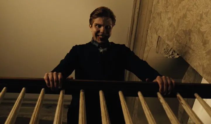 No alto de uma escada, homem jovem, branco de cabelos curtos, sorri maldosamente enquanto olha para baixo