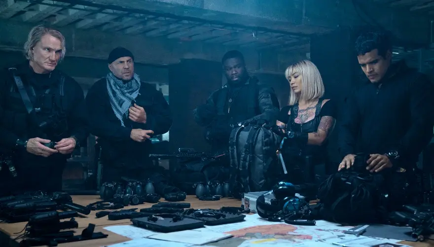 Numa sala escura, quatro homens e uma mulher loura e jovem preparam armas de fogo e equipamentos de guerra