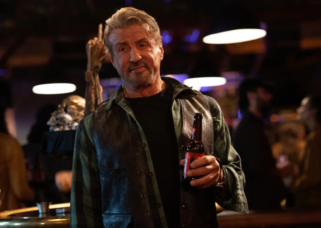 Homem de meia-idade, de cabelos grisalhos, usando blusa preta e blusão de couro, segura uma garrafa de cerveja