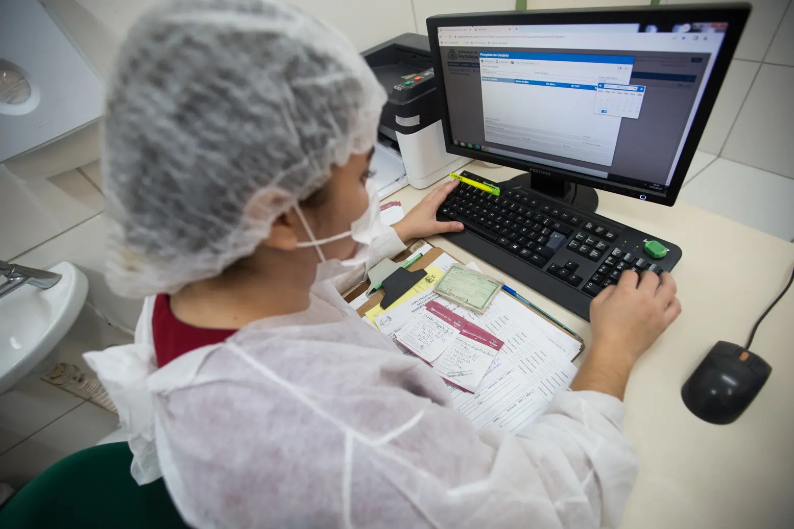 Profissional da saúde usando epi cadastra dados de paciente em computador em um posto de saúde de Fortaleza. Posto de saúde Fortaleza: saiba quando buscar, funcionamento, localizações e como ser atendido