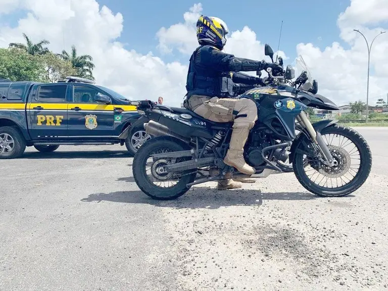 Foto de policial da PRF em moto da corporação, com viatura ao lado, em operação de Ano Novo