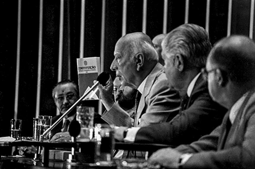 A imagem em preto e branco mostra Ulysses Guimarães ao meio, entre congressistas, segurando um exemplar da Constituição Federal de 1988 e falando ao microfone. Ele é idoso, tem cabelos brancos e é calvo. Usa vestimentas formais.